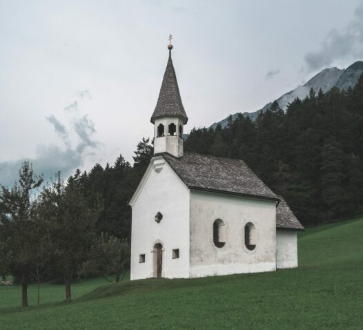 En vit kyrka med ett torn står på en grön kulle framför en skog och berg.