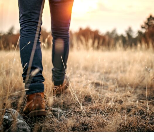 Person går i hög gräs med stövlar under en solnedgång.