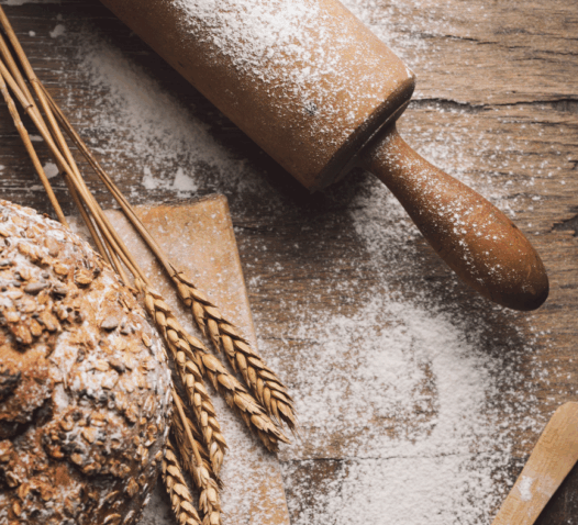 Ett nybakat bröd ligger på ett mjöligt bord bredvid ett kavel och vetestrån.