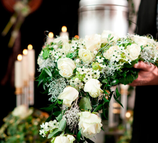 En hand håller en blomsterkrans med vita rosor och grönt lövverk, med ljus och en urna i bakgrunden.