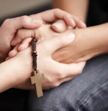 Händer som håller varandra, med ett träkors i bakgrunden.