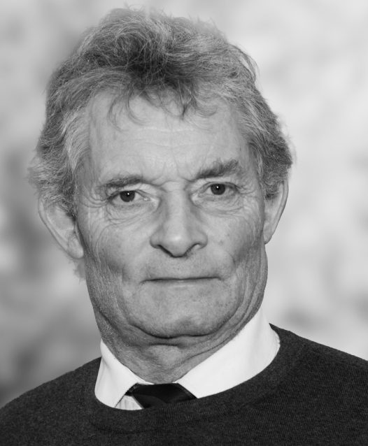 En äldre man med rufsigt hår och neutral ansiktsuttryck, klädd i skjorta och tröja, mot en suddig bakgrund.