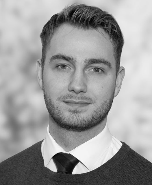 En person med kort hår och skägg, klädd i skjorta och slips, tittar mot kameran med en oskarp bakgrund.