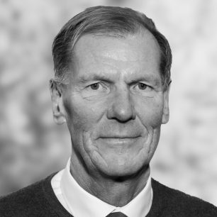 En äldre man i kavaj och slips ser in i kameran, med en neutral ansiktsuttryck och suddig bakgrund.