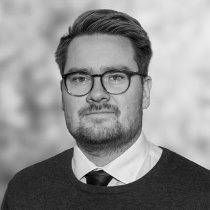 Man med glasögon och kort hår, klädd i kostym och slips, tittar mot kameran med ett neutralt ansiktsuttryck. Bakgrunden är suddig.