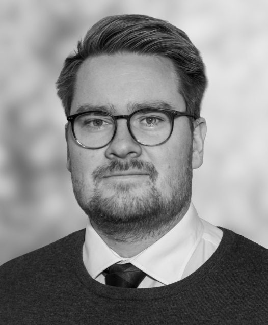 Man med glasögon och kort hår, klädd i kostym och slips, tittar mot kameran med ett neutralt ansiktsuttryck. Bakgrunden är suddig.