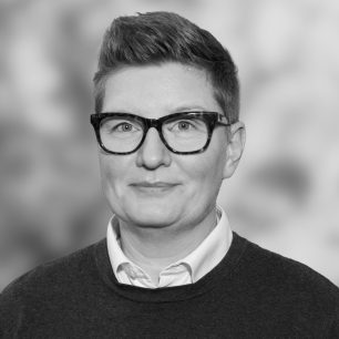 En person med glasögon och kort hår ler svagt mot kameran, klädd i en tröja och skjorta. Bakgrunden är suddig.