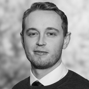 En man med kort hår och skäggstubb i svartvitt, iklädd skjorta och slips, ler lätt mot kameran.