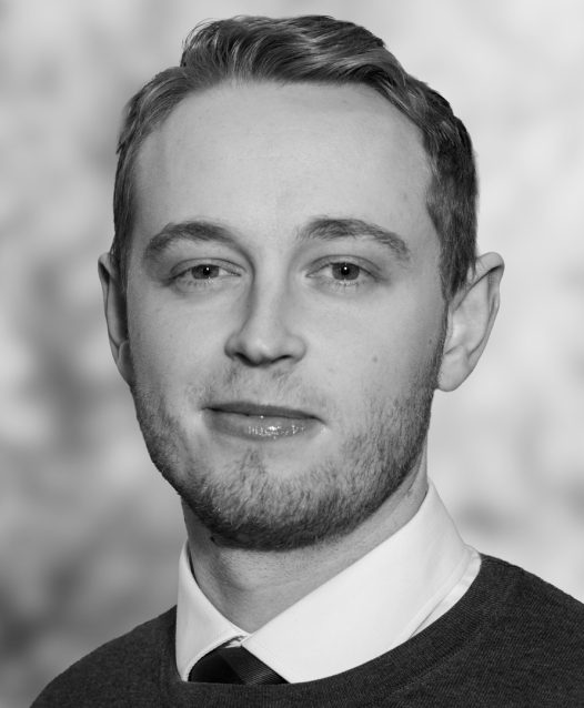 En man med kort hår och skäggstubb i svartvitt, iklädd skjorta och slips, ler lätt mot kameran.