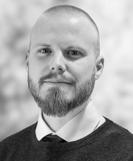 A man with a beard and shaved head wearing a sweater over a collared shirt and tie, looking at the camera against a blurred background.