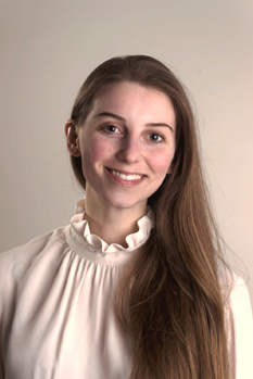 A person with long brown hair smiling, wearing a white blouse with a ruffled collar, set against a neutral background.