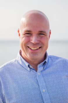 A bald man smiling while wearing a light blue button-up shirt, standing in front of a blurred outdoor background.