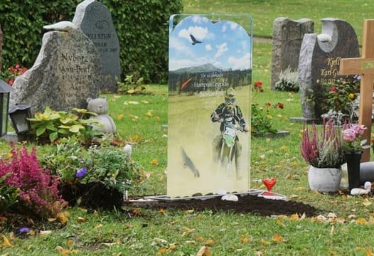 En kyrkogård med flera gravstenar. En gravsten har en bild av en motorcyklist och texten "Vi saknar dig mycket, du finns i våra hjärtan alltid." Runt gravarna finns blommor och dekorationer.
