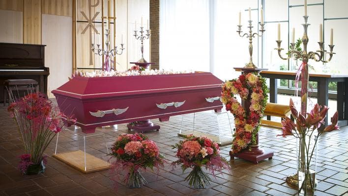 En rosa kista omgiven av blomsterarrangemang och ljusstakar i en kyrka.