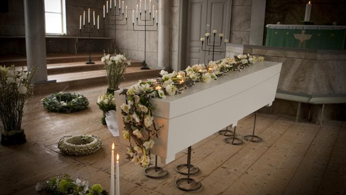 Vit kista dekorerad med blommor och ljus i en kyrka omgiven av kransar och tända ljus.