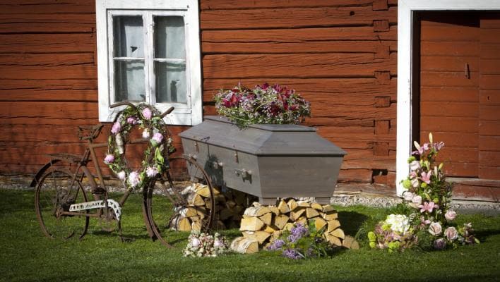 En träkista dekorerad med blommor står på vedträn framför en röd träbyggnad.
