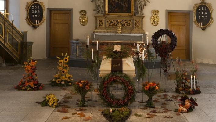 Kista dekorerad med blommor och ljus i en kyrka, omgiven av kransar och höstlöv.