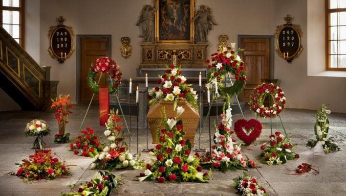 Kista omgiven av blomsterarrangemang i en kyrka, med tända ljus och en tavla i bakgrunden.