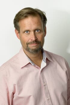 A person with short hair and a beard wearing a light pink button-up shirt looks directly at the camera against a plain white background.