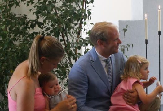 En man i kostym och en kvinna i rosa klänning sitter med två små barn i ett rum med tända ljus och gröna växter.