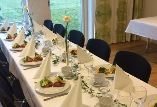 Ett långbord är dukat med vita dukar, tallrikar med mat, vikta servetter och glas. Bordet är dekorerat med blommor och gröna växter, och står i ett rum med fönster mot en gräsmatta.