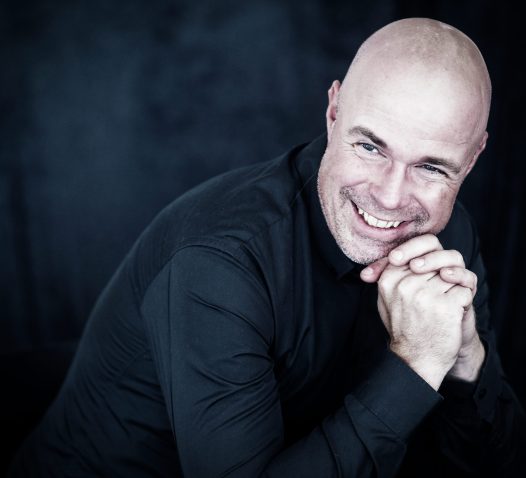 A person in a black shirt smiles warmly with hands clasped under their chin against a dark background.