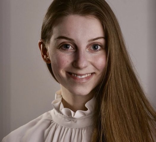 A person with long hair and a white ruffled blouse smiles at the camera against a plain background.
