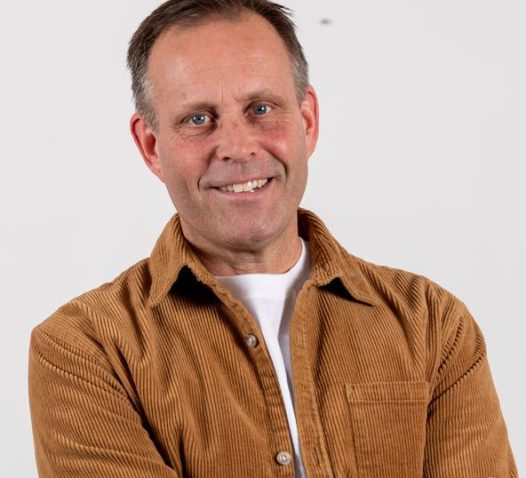 A person wearing a brown corduroy shirt smiles with arms crossed in front of a plain background.